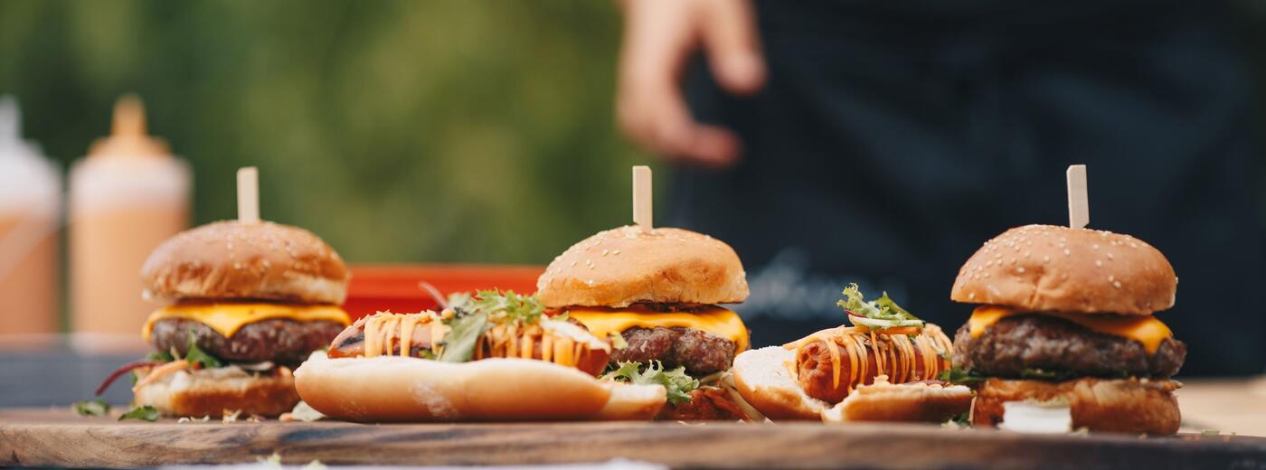 Picnic table with blue gingham tablecloth, and a cutting boards with hamburgers and hotdogs