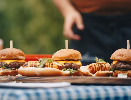 Picnic table with blue gingham tablecloth, and a cutting boards with hamburgers and hotdogs